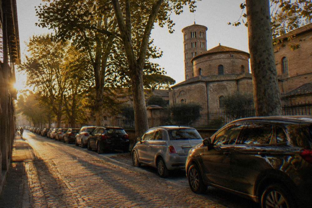 Cars parked in a row along a tree-lined street outside a historic centre