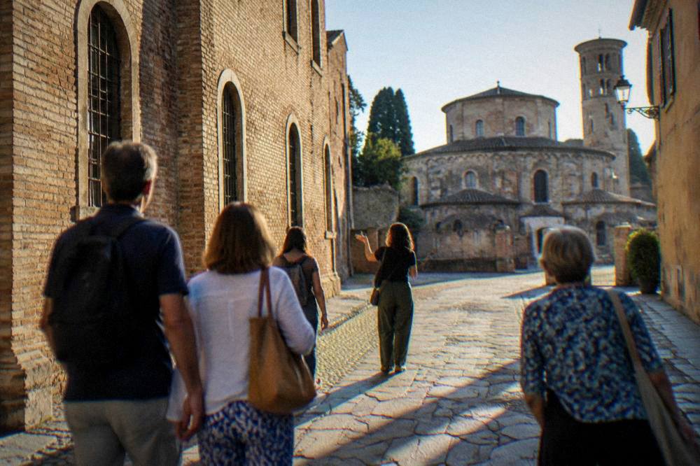 A small group of visitors following a guide along a sunlit street of old brick facades