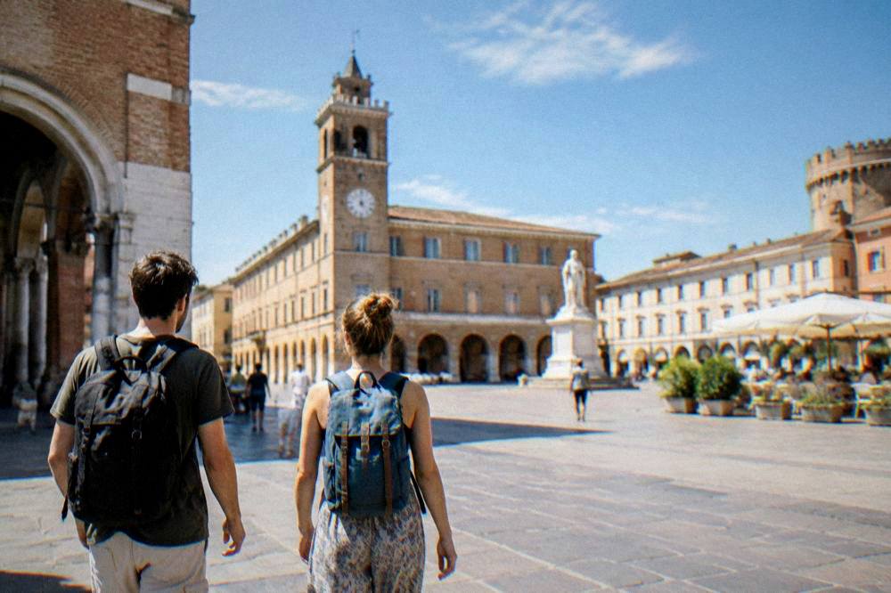 Two people with backpacks walking through a sunlit Italian piazza