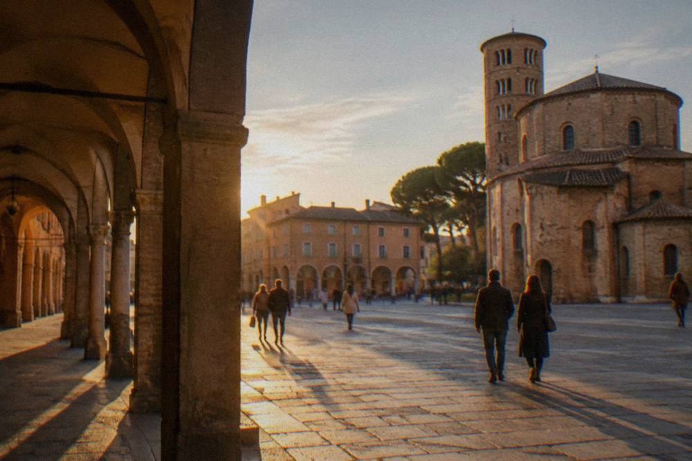 Visitors walking across a sunlit historic square lined with arcaded buildings