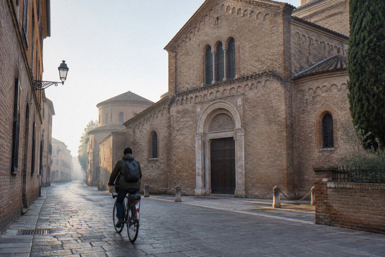 A quiet Ravenna street lined with brick buildings and a cyclist passing an old church facade