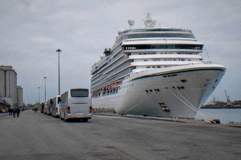 A cruise ship docked at an industrial port quay with shuttle buses waiting nearby